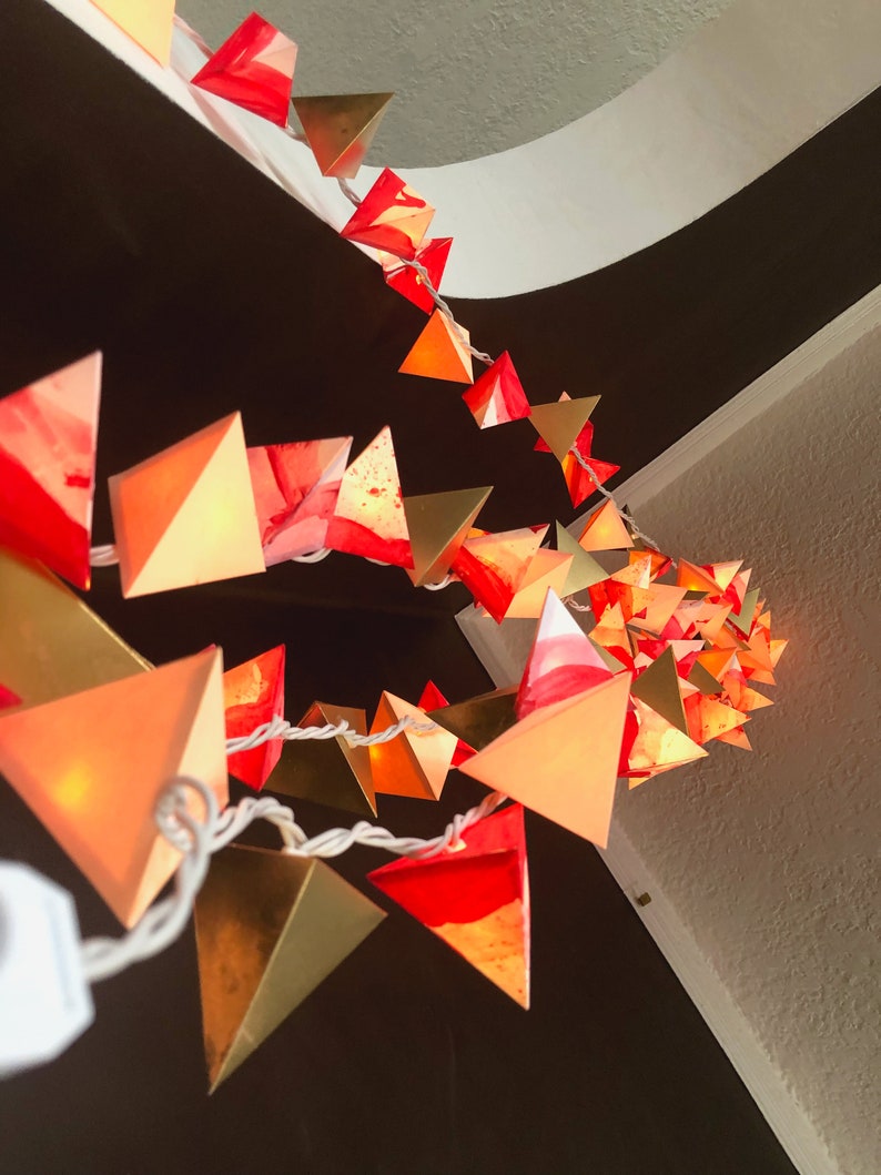 May include: A string of lights with red and gold triangular paper lanterns. The lanterns are lit and glowing, creating a warm and festive atmosphere.