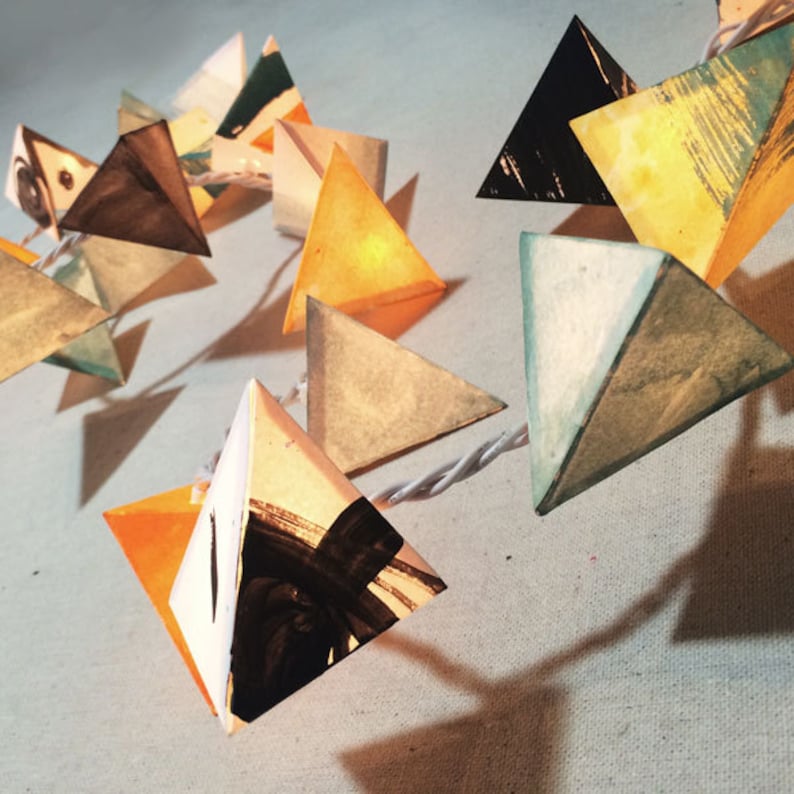 May include: A string of triangle-shaped paper lanterns with a white wire. The lanterns are painted in various colors, including yellow, orange, blue, green, and black.