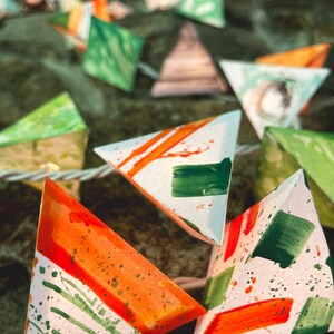May include: A string of triangular paper lanterns with a white background and green and orange watercolor designs. The lanterns are strung together with white string.