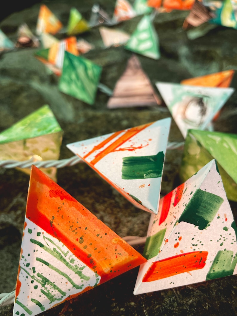 May include: A string of triangular paper lanterns with a white background and green and orange watercolor designs. The lanterns are strung together with white string.