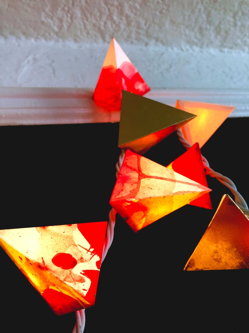 May include: A string of pyramid-shaped lights with a red, gold, and orange color scheme. The lights are made of paper and have a watercolor-like design.