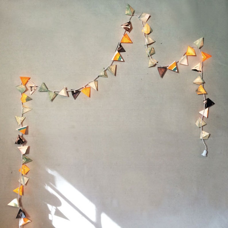 May include: A string of triangle-shaped paper lanterns in shades of yellow, green, and brown, strung on a white cord. The lanterns are lit and create a warm, festive atmosphere.