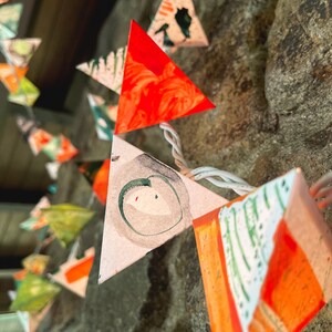 May include: A string of triangular paper lanterns with a white background and orange, green, and black painted designs. The lanterns are strung together with white wire.