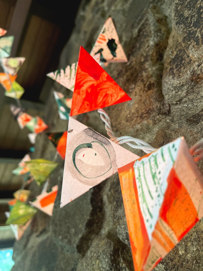 May include: A string of triangular paper lanterns with a white background and orange, green, and black painted designs. The lanterns are strung together with white wire.