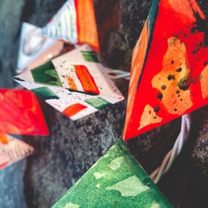 May include: A string of colorful paper lanterns with green, orange, and red geometric patterns. The lanterns are strung together with white cord and are hanging from a dark background.