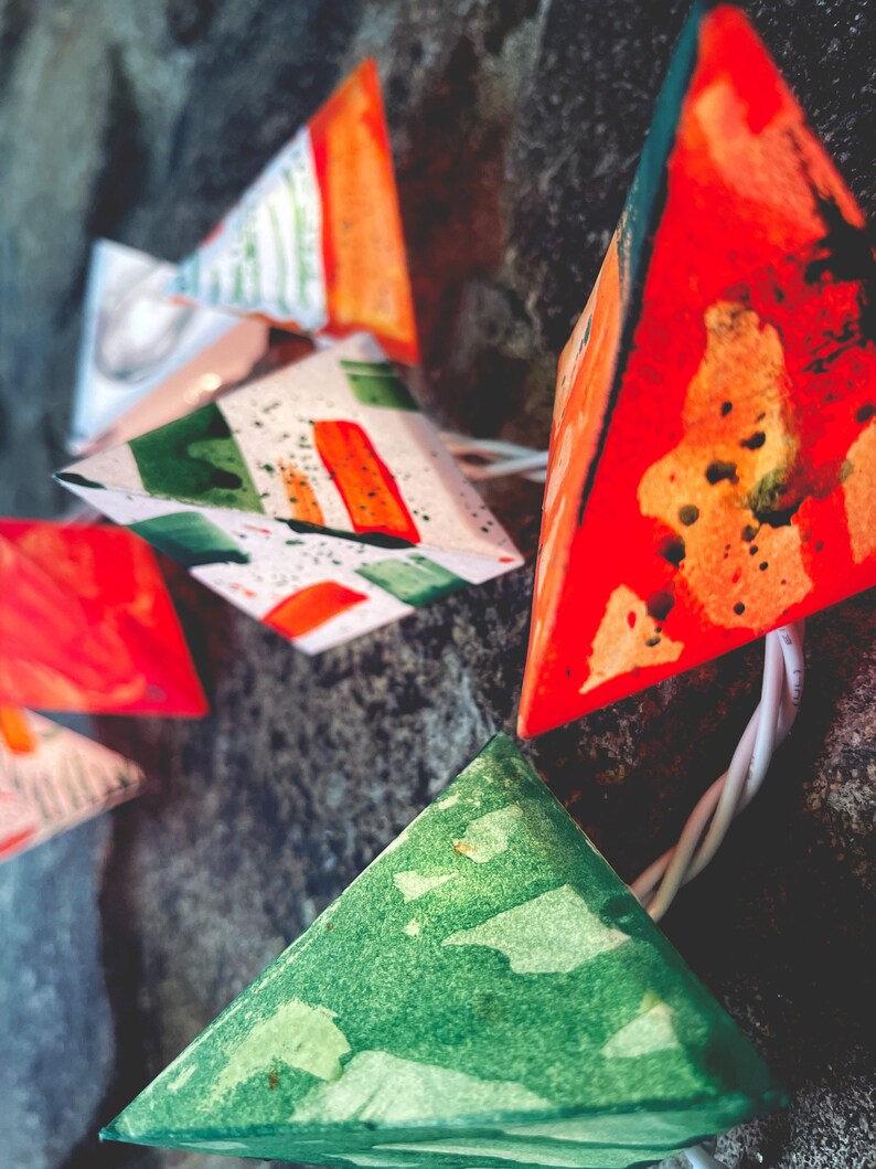 May include: A string of colorful paper lanterns with green, orange, and red geometric patterns. The lanterns are strung together with white cord and are hanging from a dark background.