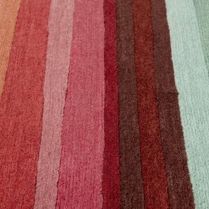 May include: A rectangular area rug featuring a series of vertical stripes in a gradient of colors including orange, various shades of red, deep brown, and light mint green. The rug has a textured, tufted pile surface with a consistent weave pattern.