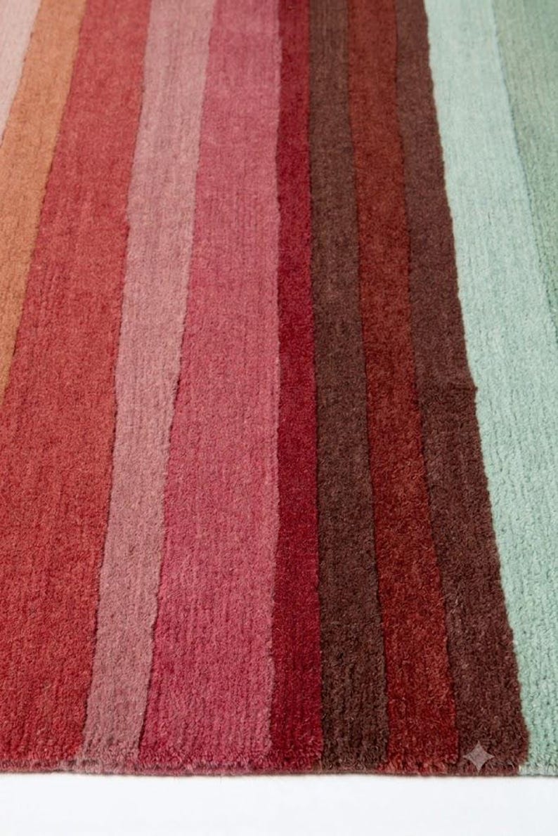 May include: A rectangular area rug featuring a series of vertical stripes in a gradient of colors including orange, various shades of red, deep brown, and light mint green. The rug has a textured, tufted pile surface with a consistent weave pattern.