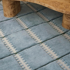 May include: A light blue area rug featuring a repeating grid pattern of rectangular sections separated by white vertical bands with raised tufted textures. The rug has a soft, plush pile and is positioned beneath a light-colored wooden table.
