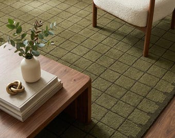 Puede incluir: Una alfombra de color verde oliva con un patrón de cuadrícula repetitivo de cuadrados de color verde oscuro. La alfombra tiene una superficie texturizada y copetuda, extendiéndose por el suelo bajo un sillón blanco y una mesa de madera.