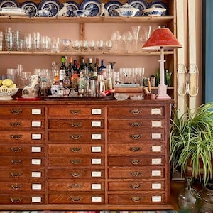 May include: A large wooden apothecary cabinet with many small drawers, topped with open shelving holding blue and white ceramic plates, glassware, and a red lamp. A striped rug lies in front, and a green plant sits in a glass vase nearby.
