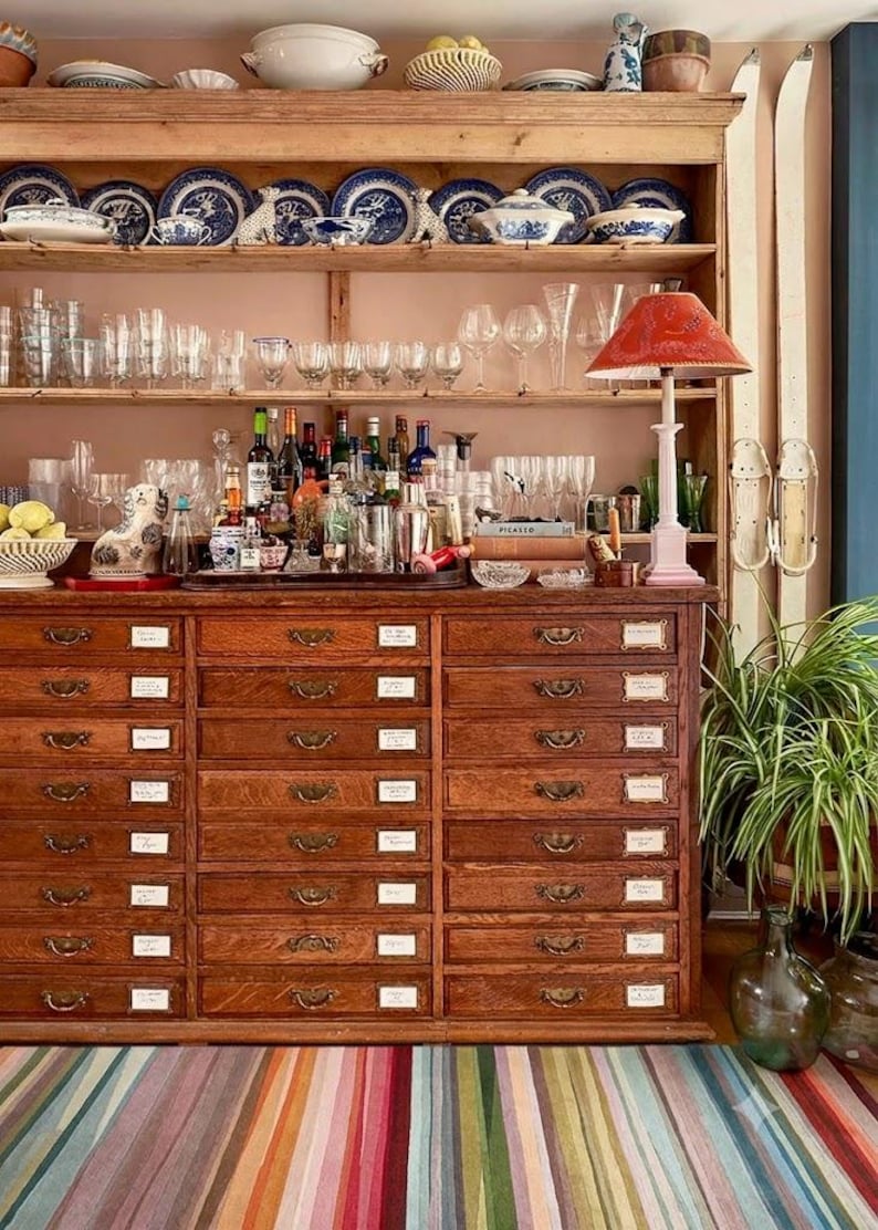 May include: A large wooden apothecary cabinet with many small drawers, topped with open shelving holding blue and white ceramic plates, glassware, and a red lamp. A striped rug lies in front, and a green plant sits in a glass vase nearby.