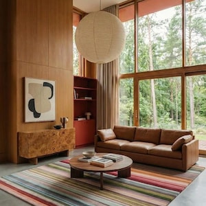 May include: A large rectangular area rug featuring horizontal stripes in various colors including red, green, blue, and beige. The rug is placed on a concrete floor in a living room with a brown leather sofa, a round wooden coffee table, and wood walls.