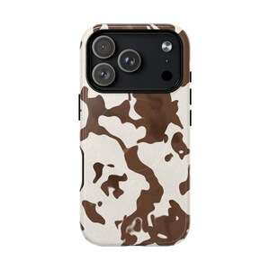 May include: A smartphone case featuring a brown and white cow print pattern. The case has a black camera module cutout at the top, revealing three circular camera lenses and a flash. The design covers the back of the device with irregular patches.