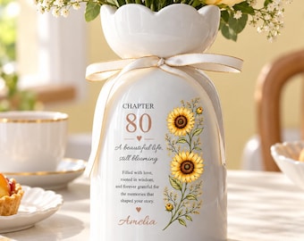 May include: A white ceramic vase with a scalloped rim and a cream ribbon tied around the neck. The vase features the text "CHAPTER 80 A beautiful life, still blooming" with a sunflower illustration and the name "Amelia" in script.