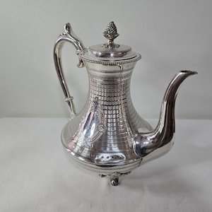 Peut inclure: Une théière en argent poli présentant un motif festonné texturé sur le corps, une anse incurvée, un long bec verseur et un couvercle surmonté d'un petit fleuron. La base repose sur trois petits pieds et un cartouche ovale lisse est centré.