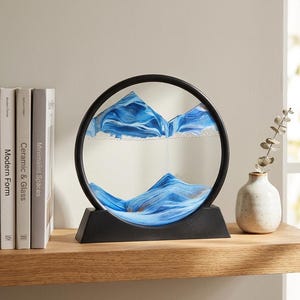 May include: A circular sand art frame with a black border and base, containing layers of blue, white, and gold sand that form mountain-like shapes within the glass. The sand is suspended in a clear liquid, creating distinct, flowing landscape patterns.