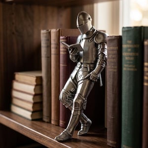 Può includere: Una statuetta in metallo color argento raffigurante un cavaliere in armatura completa in piedi che legge un libro aperto. Il cavaliere è raffigurato con elmo, spallacci e schinieri, appoggiato leggermente mentre tiene il libro con entrambe le mani.