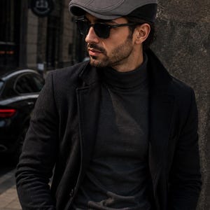 May include: A grey flat cap featuring a structured crown, a short stiff brim, and two small metal eyelets on the side. The cap is constructed with a smooth fabric finish and a dark band detail along the lower edge, providing a clean and simple profile.