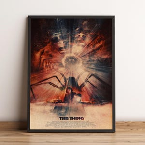 Op de afbeelding: Een ingelijste filmposter voor The Thing met een centraal figuur in een besneeuwd landschap onder fel licht, omringd door donkere, vervormde gezichten en het silhouet van een spinachtig wezen, uitgevoerd in tinten rood, zwart en wit.