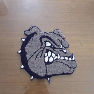 Puede incluir: Un parche textil con mechones en forma de cabeza de bulldog de perfil. El diseño presenta una cara gris con contornos de color morado oscuro, dientes blancos y un collar de púas de color morado oscuro con púas blancas. Textura densa y lanuda.