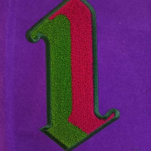 May include: A patch in the shape of the number one, featuring a split design with a green left side and a pink right side. The patch has a dark green border and is attached to a solid purple fabric background with a textured, fuzzy surface.