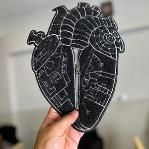 Peut inclure: Un écusson en forme de cœur noir avec un contour brodé blanc et des motifs géométriques. Une fermeture éclair en métal argenté descend verticalement au centre du cœur, divisant la surface en tissu noir texturé en deux sections.