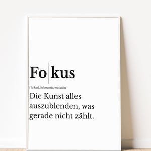 Könnte beinhalten: Ein weiß gerahmter Kunstdruck mit dem schwarzen Text 'Fokus', gefolgt von der phonetischen Aussprache, dem Wort 'Substantiv, maskulin' und der Definition 'Die Kunst alles auszublenden, was gerade nicht zählt.'