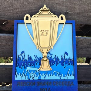 May include: A layered wooden wall plaque featuring a gold-colored trophy with the number 27 in the center. Below the trophy is a silhouette of a cheering crowd in shades of blue. The text "ЛЕВСКИ ЗНАЧИ СВОБОДА" and "1914" is engraved at the bottom.