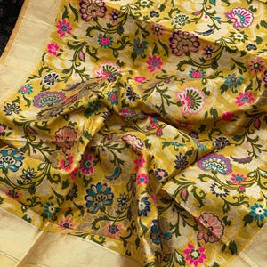 Könnte beinhalten: Ein senfgelber Stoff mit einem dichten, gewebten Blumenmuster in den Farben Pink, Petrol, Dunkelgrün und Creme. Das Textil weist entlang der Kante eine breite, einfarbige, metallisch goldene Bordüre mit einer fein strukturierten Oberfläche auf.