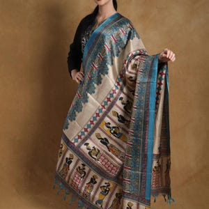 May include: A beige silk dupatta featuring intricate hand-painted figures in black, yellow, and red, framed by a wide teal border with ornate floral patterns. The fabric is finished with teal tassels along the edges, showcasing a traditional aesthetic.