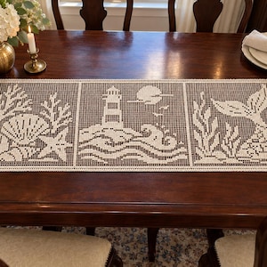 Puede incluir: Un camino de mesa rectangular de ganchillo en color blanco roto con tres paneles distintos. El panel izquierdo muestra conchas y coral, el central un faro con sol y olas, y el derecho representa una cola de sirena con algas marinas.