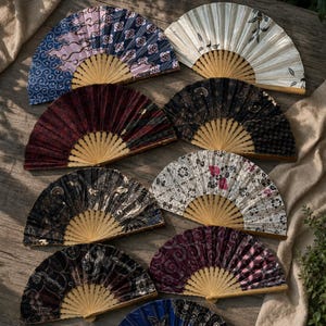 Peut inclure: Une collection de neuf éventails pliants avec des baleines en bambou et des tissus aux motifs variés, incluant des imprimés floraux, géométriques et de style batik dans des tons de bleu, blanc, noir, rouge et violet, disposés en éventail.