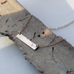 May include: A gold bar necklace with a delicate chain and a personalized initial "K" engraved on the bar.