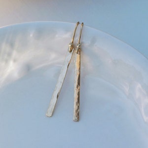 May include: A pair of gold hammered bar earrings with a simple design. The earrings are made of a smooth, polished metal and have a hammered texture.