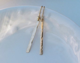 May include: A pair of gold hammered bar earrings with a simple design. The earrings are made of a smooth, polished metal and have a hammered texture.
