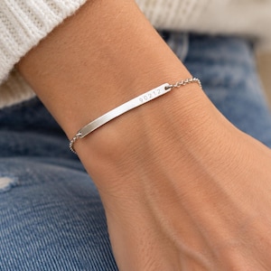 May include: A silver-toned metal bar bracelet featuring a delicate chain and a flat, rectangular center plate. The plate is engraved with the numerical text 90212. The bracelet is designed with a simple, clean aesthetic and a secure clasp mechanism.