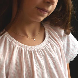 May include: A delicate gold-toned chain necklace featuring a small, simple cross pendant. The necklace rests against the neck, showcasing a thin, polished metal link design that connects to a tiny, centered cross charm with clean, straight edges.