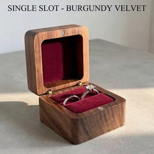 May include: A square wooden ring box with a hinged lid, featuring a dark brown wood grain exterior and a deep burgundy velvet interior lining. The text SINGLE SLOT - BURGUNDY VELVET is printed above the box, which holds two silver rings.
