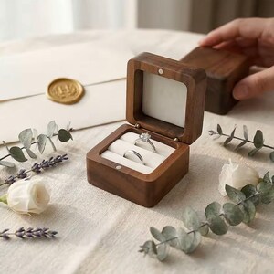 May include: A square dark brown wooden ring box with a hinged lid and a white interior insert holding two silver-toned rings, one featuring a prominent round stone setting and the other a plain band, displayed in an open position.