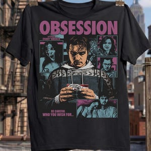 Könnte beinhalten: Ein schwarzes Kurzarm-T-Shirt mit einem Grafikdruck, der den Text OBSESSION, A FILM BY CURRY BARKER und BE CAREFUL WHO YOU WISH FOR enthält. Das Design zeigt mehrere illustrierte Felder mit Personen und eine zentrale Figur, die eine kleine Karte hält.