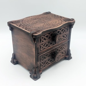 Peut inclure: Un coffret en bois brun foncé doté de deux tiroirs, orné de motifs de nœuds sculptés sur la façade et d'un dessin d'arbre circulaire sur le couvercle. Chaque tiroir possède un bouton rond affichant un symbole triskèle à triple spirale.