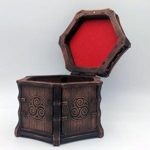 Peut inclure: Une boîte hexagonale en bois avec une finition brun foncé et un couvercle à charnière. L'extérieur présente des symboles triskèles sculptés sur chaque panneau latéral. L'intérieur du couvercle est doublé d'un tissu rouge vif contrastant.