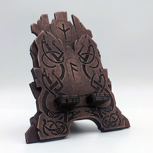 Könnte beinhalten: Ein dunkelbrauner Handyhalter aus Holz mit detaillierten, laser-gravierten Knotenmustern im nordischen Stil und zwei zentralen Runensymbolen. Der Halter hat eine gezackte, steinartige Oberkante und zwei kleine Vorsprünge als Stütze.