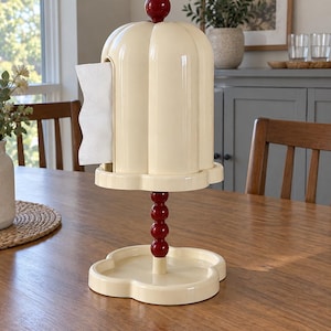 Puede incluir: Un soporte para papel de cocina de color crema con una cubierta en forma de cúpula y un remate esférico rojo. El poste central presenta una pila de cuatro esferas rojas y la base tiene forma de trébol de cuatro lóbulos. Se ve una hoja blanca.