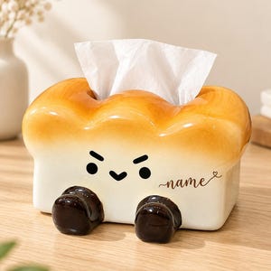 May include: A ceramic tissue box cover shaped like a loaf of bread with a golden-brown crust top and a cream-colored base. It features a cartoon face with an angry expression, two dark brown feet, and the word name printed in cursive script.