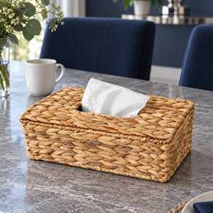 May include: A rectangular tissue box cover crafted from woven natural seagrass in a light tan color. The box features a braided texture and a central opening at the top, through which a single white paper tissue is pulled upward for easy access.