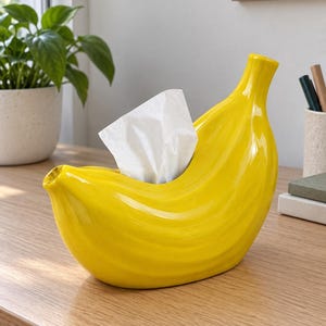 May include: A bright yellow ceramic tissue box cover shaped like a curved banana. The glossy, textured surface mimics the natural ridges of a banana peel. A single white facial tissue is pulled through the central opening at the top.