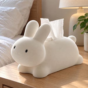 Op de afbeelding: Een witte tissueboxhouder in de vorm van een konijn met een gladde, matte afwerking. Het ontwerp heeft twee rechtopstaande oren, een ronde kop met twee kleine zwarte ogen en een klein mondje. Een witte tissue steekt uit de opening.