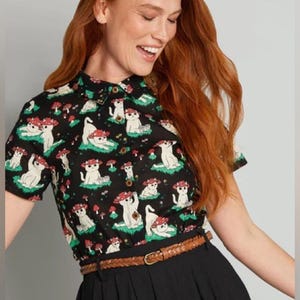 May include: A black short-sleeved button-down shirt featuring a repeating pattern of white cats wearing red mushroom caps, paired with a brown braided belt over black trousers. The shirt has a classic collar and dark brown buttons.